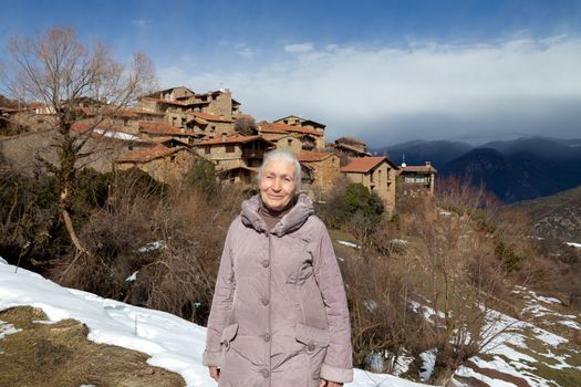 An elderly woman enjoys a landscape in a highland authentic Catalan village with stone houses while walking along a mountain path covered with melting snow.
