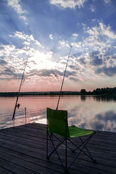 Folding fishing chair and two fishing rods on a wooden pontoon on the lake at sunset