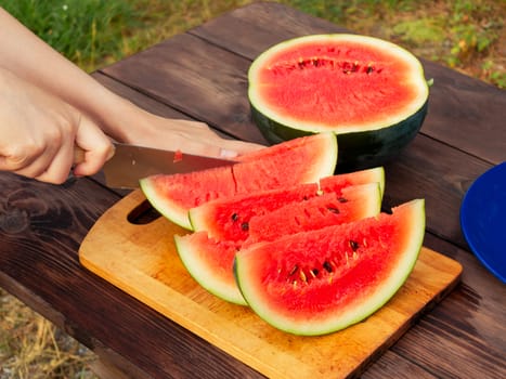Women's hands cut with a knife into slices of ripe watermelon on a wooden table.