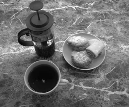 A cup of black tea, a teapot and cookies on a glass saucer are lying on a marble table. Black and white shot.