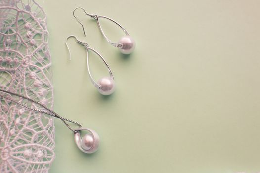 Fashion women's silver accessories - earrings and pendant with pearls on light background with rich openwork lace, top view, flat, space for text.