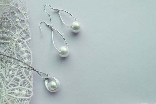 Fashion women's silver accessories - earrings and pendant with pearls on light background with rich openwork lace, top view, flat, space for text.