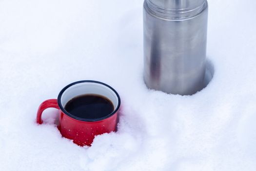 Red metal cup with coffee poured from a thermos on pure white snow in the forest while walking.