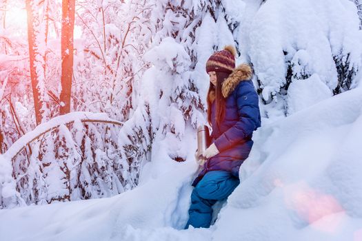 girl with a thermos in her hands is walking in the snowy winter forest.