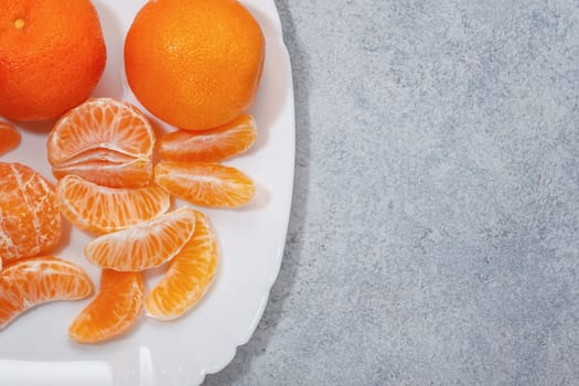 Several whole and peeled ripe tangerines on a white plate on gray background with space for text, flat lay, top view.