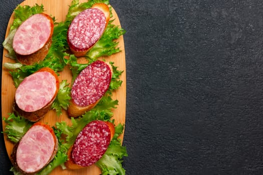 Several sandwiches with sausage and salami and sauce on a black board, flat lay background with copyspace.