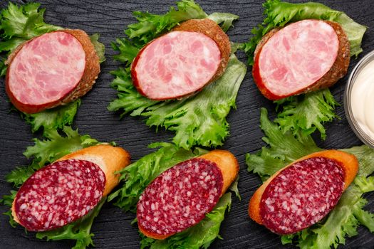 Several sandwiches with sausage and salami and sauce on a black board, flat lay.