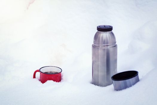 Red metal cup with coffee poured from a thermos on pure white snow in the forest while walking.