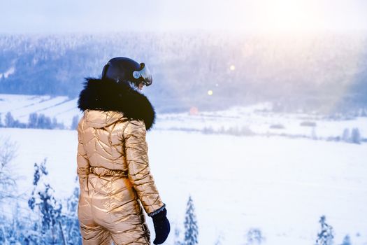 young woman in a golden ski overalls and helmet stands on top of a slope and looks into the distance.