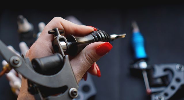 Female hand with red nails holding tattoo machine. Hand without gloves - practice and training tattoo. Dark background