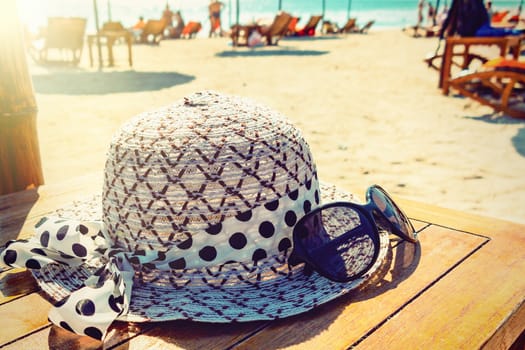 Women's hat and sunglasses lie on a wooden table on a sunny sandy beach by the sea.