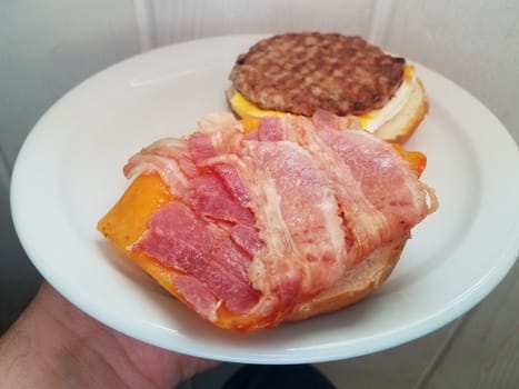 hand holding sausage and egg and bacon and cheese sandwich on white plate