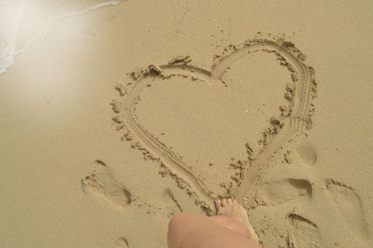 Hello summer, women's feet draw a heart sign on the wet sand while standing on the sandy beach. Concept of rest, relaxation, top view.