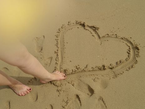 Hello summer, women's feet draw a heart sign on the wet sand while standing on the sandy beach. Concept of rest, relaxation, top view.