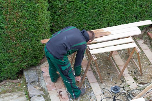 A man holds a paintbrush in hand and strokes paint color on wooden furniture. Concept renovation garden furniture.