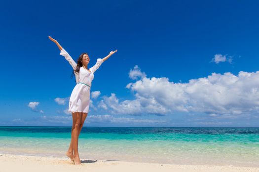 Woman in white dress posing in tropical sea beach with arms raised