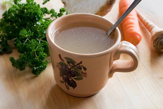 Chicken bone broth in a mug, with parsley, carrot and celery root in the background