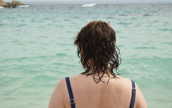 View of the back of an adult woman in a swimsuit with wet hair, standing on the beach and admiring the sun glare on the sea,