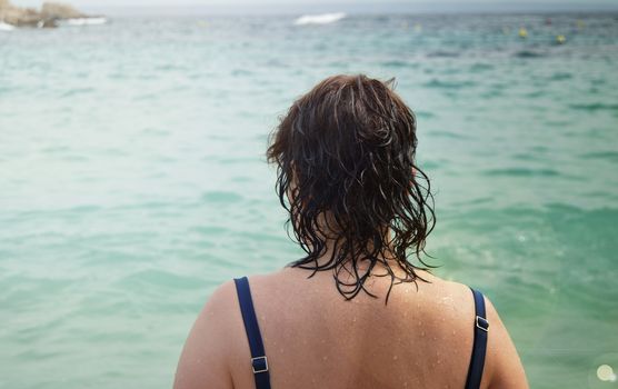 View of the back of an adult woman in a swimsuit with wet hair, standing on the beach and admiring the sun glare on the sea.