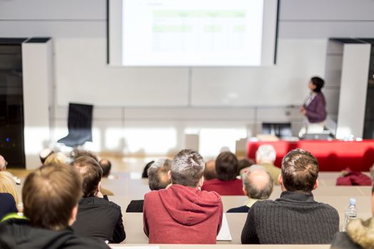 Female speaker giving academic presentation in lecture hall at university workshop. Audience in conference hall. Rear view of unrecognized participant in audience. Scientific conference event.