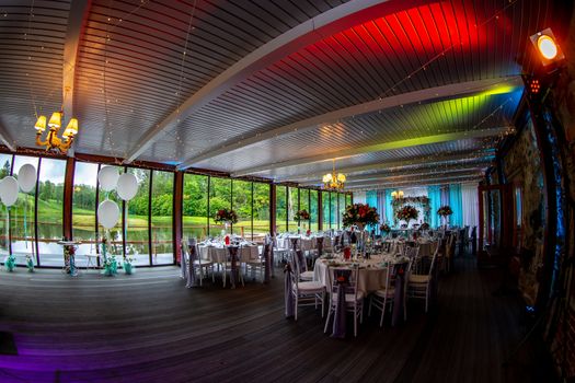 Luxury decorated place for wedding reception catering in restaurant next to the pond. Guest tables in luxury decorated wedding banquet room. Festive decorations and items for food, arranged on the tables.