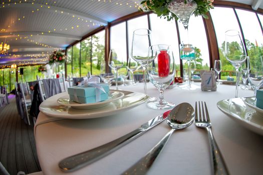 Table setting with handmade gift box on plate, bouquet of flowers, glasses, forks, spoons and knives for wedding reception. Guest tables in luxury decorated wedding banquet room. Shot with fisheye lens. 