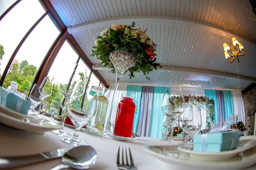 Table setting with handmade gift box on plate, bouquet of flowers, glasses, forks, spoons and knives for wedding reception. Guest table in luxury decorated wedding banquet room. Shot with fisheye lens. 