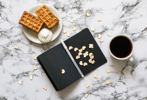 black sheets notebook with white pen on marble background and waffles, coffee