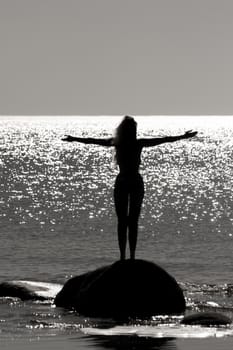 Photo of a girl standing on a stone with her arms spread apart. By the sea. Against the shimmering glare of the sun on the surface of the sea. Counter shooting. Black and white photo.