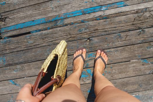 Top view, photo of legs in beach flip-flops and with a straw bag in hand on a wooden old floor. Photos on vacation, beach, summer.