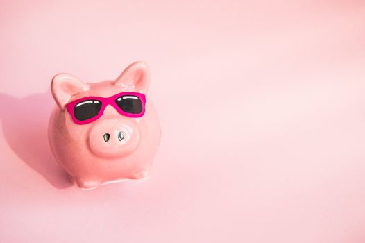 Funny piggy Bank in sunglasses on pink background, sunlight, copy space, money saving concept for summer vacation.