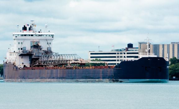 A cargo ship with metal aboard sails along the River Detritus past city buildings