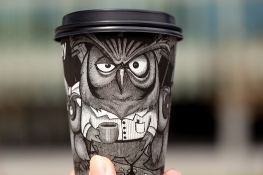 Black paper disposable coffee cup with a drawn owl in hand on a blurred background outdoor