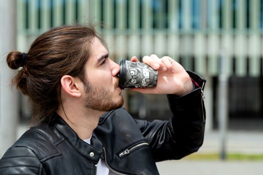 Portrait of a handsome fashionable hipster man in a black leather jacket drinking tea or coffee from a black takeaway paper cup outside.