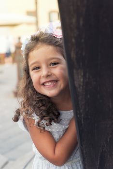 little girl smiles playfully while hiding, wearing a blue dress and a flower headband