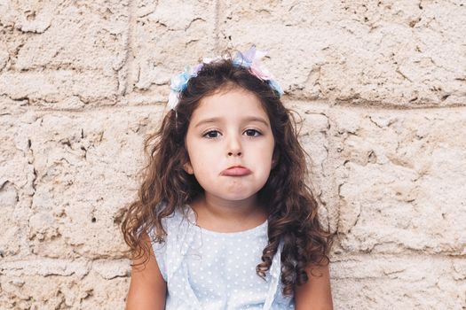 little girl making teasing with sad face, is in front of a stone wall and wears a blue dress and a flower headband