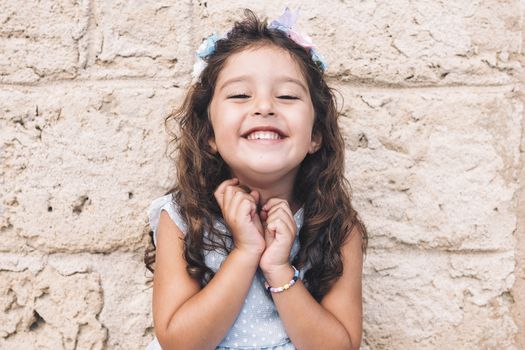 little girl smiles funny, is in front of a stone wall and wears a blue dress and a flower headband