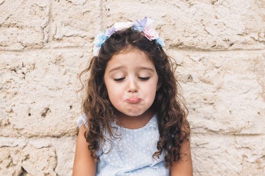little girl making fun with sad face, is in front of a stone wall and wears a blue dress and a flower headband