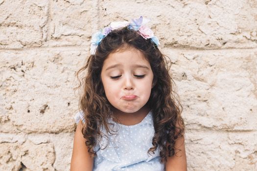 little girl making fun with sad face, is in front of a stone wall and wears a blue dress and a flower headband