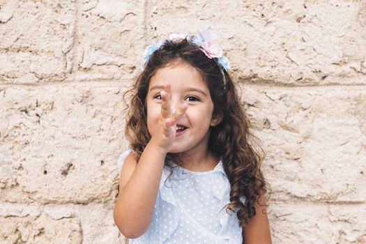 little girl making fun with her hand on her nose, is in front of a stone wall and wears a blue dress and a flower headband