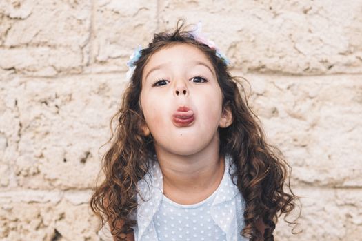 Teasing little girl sticking out her tongue, is in front of a stone wall and wears a blue dress and a flower headband