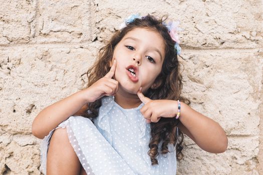 little girl making fun with hands and face, is in front of a stone wall and wears a blue dress and a flower headband