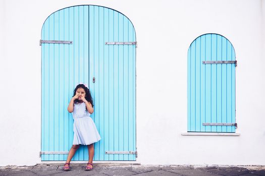 funny little girl making fun with her eyes, she is in front of a white wall with a door and a turquoise wooden window