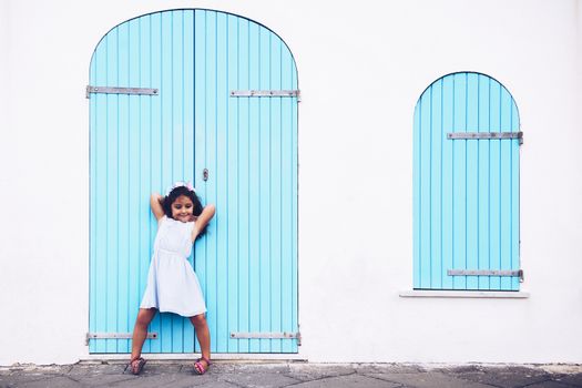 funny little girl making fun, she is in front of a white wall with a door and a turquoise wooden window
