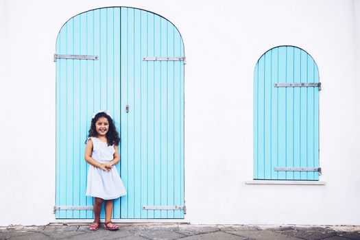 little girl smiles funny, she is in front of a white wall with a door and a turquoise wooden window