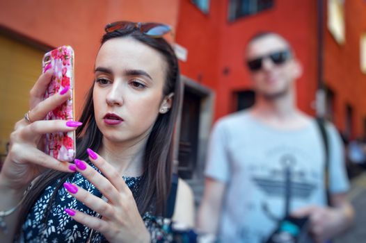 Woman looks into the phone and walks down the street with a man behind. Model wearing stylish sunglasses. City lifestyle. Female fashion concept.