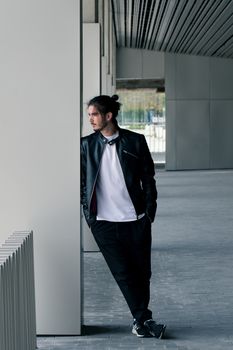 Bearded handsome young man in a black leather jacket with a stylish haircut stands and leans on the wall, looking away.