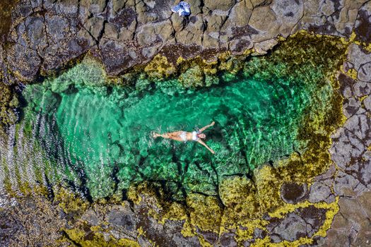 A woman relaxes in a beautiful seaside rock pool with sandy bottom and edged in a yellow seaweed called Neptunes necklace, Hormosira banksia