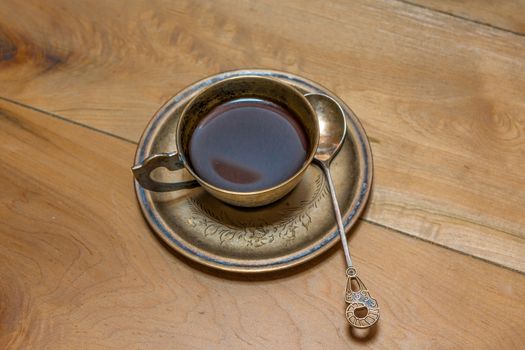 A cup with black coffee, a saucer and a spoon made of bronze stand on an oak table