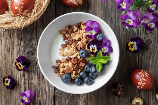 Breakfast cereals with yogurt, blueberries and edible pansy flowers, with Easter eggs dyed with onion peels in the background